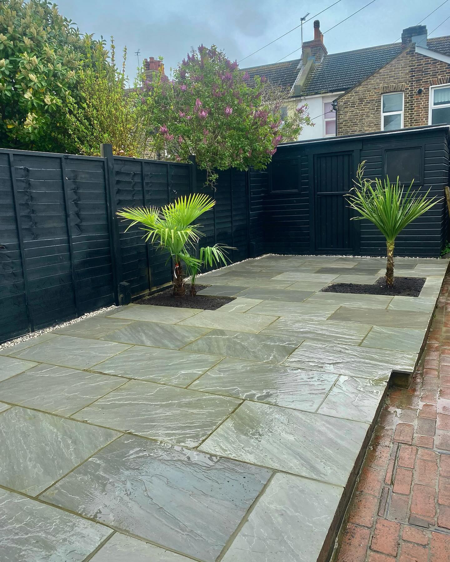 A grey slab courtyard with a black painted fence and palms in planters